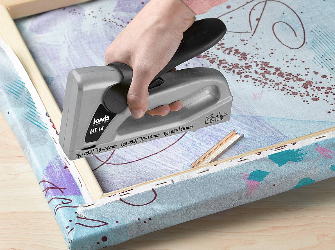 Quelqu'un fixe une toile sur un cadre en bois à l'aide d'un agrafeur noir et gris.