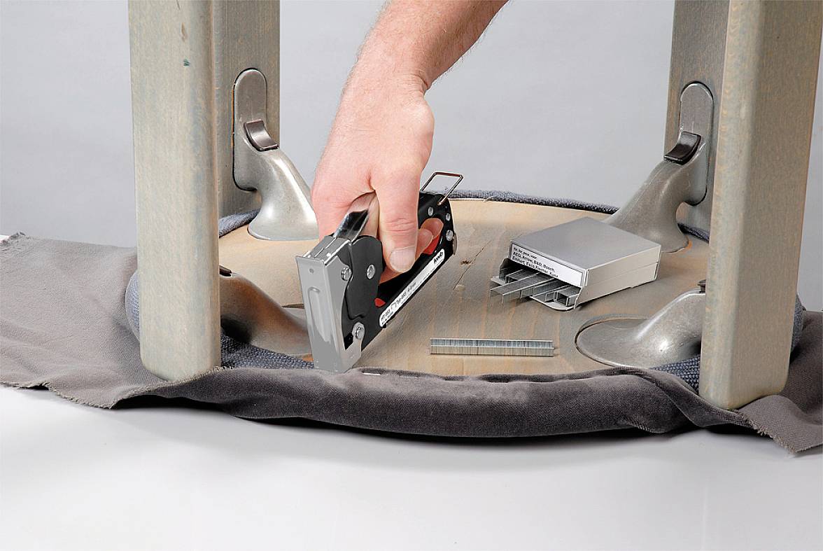 Une main utilise un pistolet à agrafes pour fixer le tissu sur l'assise d'une chaise retournée. Des agrafes sont visibles à côté.