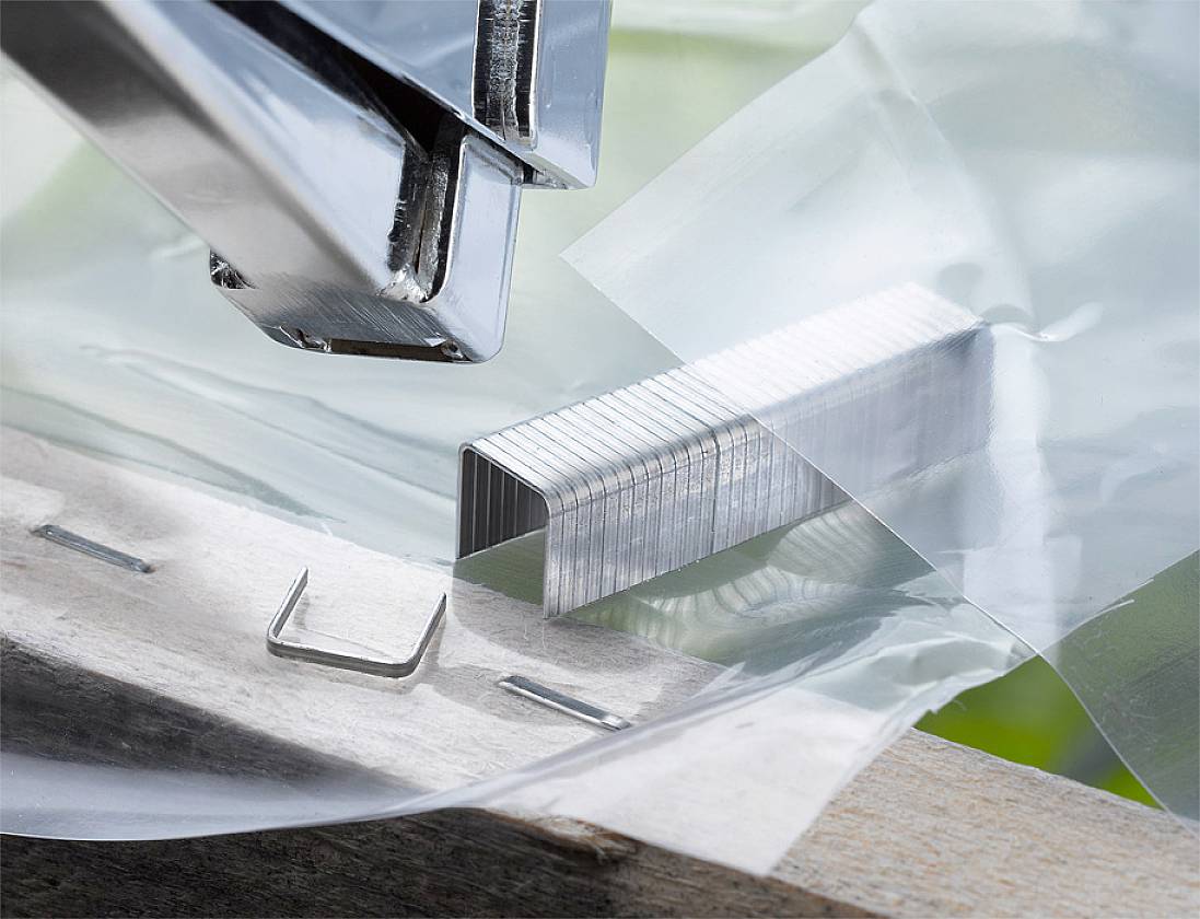 Un agrafeur argenté traverse une feuille de plastique. Un tas d'agrafes est posé à côté.