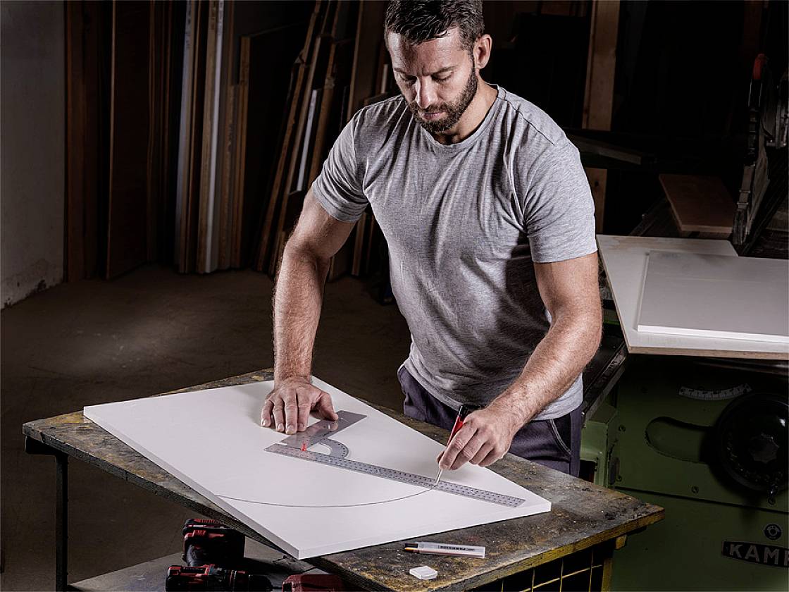 Un homme portant un t-shirt gris dessine une courbe sur un tableau blanc à l'aide d'une règle et d'un crayon dans un atelier.