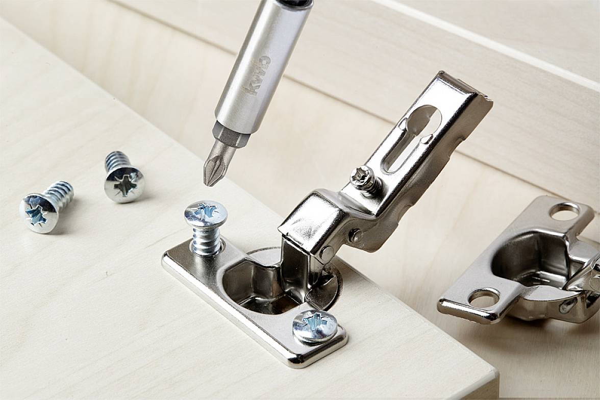 Un tournevis enfonce une vis dans une charnière sur une planche en bois. Deux autres vis sont posées à côté de la charnière.