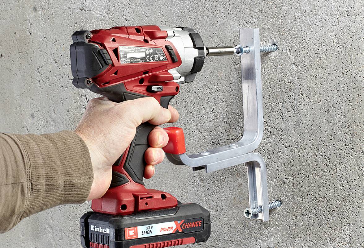 Une main tient un tournevis-perceuse rouge sans fil qui visse un support métallique dans un mur en béton.