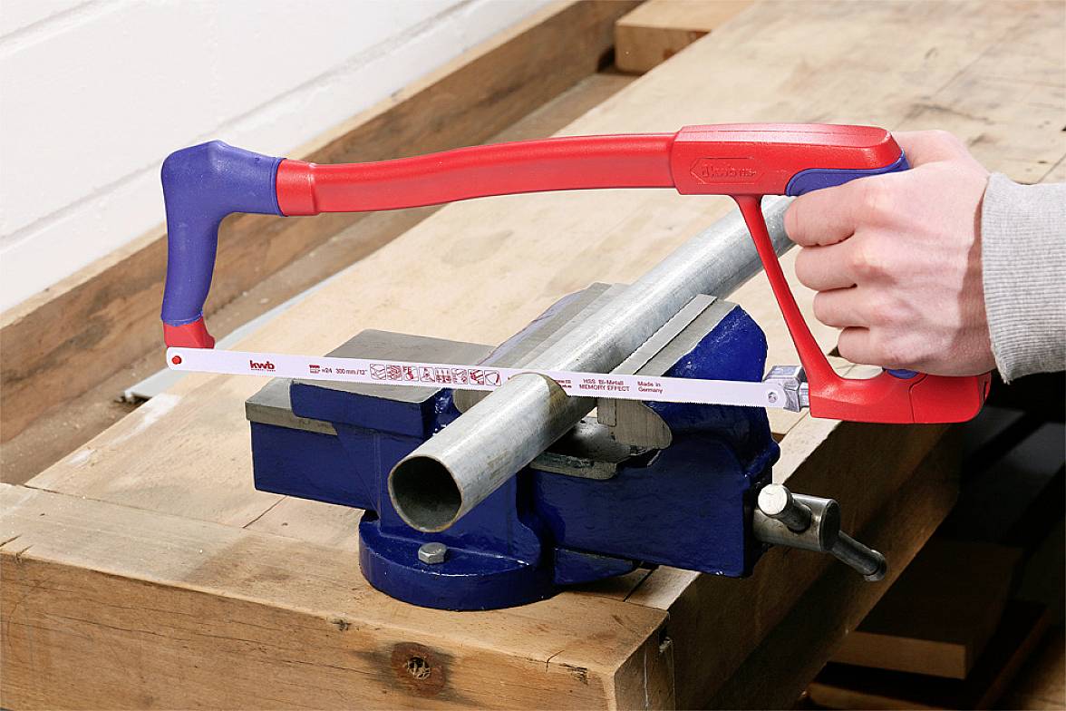 Une personne scie un tuyau métallique avec une scie à métaux, fixé dans un étau de établi bleu.