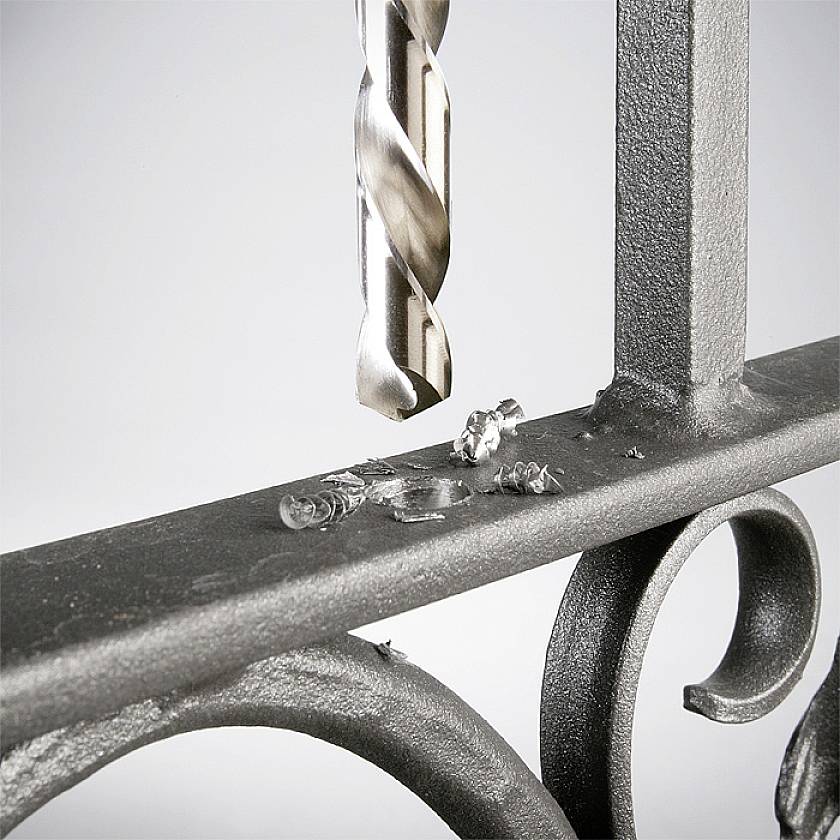 Un foret perce une rambarde métallique, générant des copeaux de métal ; le focus est mis sur le processus de perçage et les copeaux.