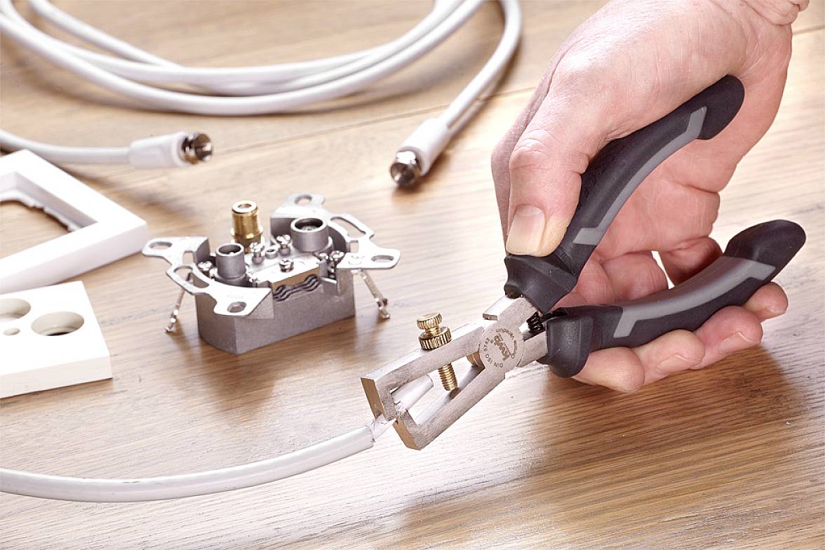 Une main utilise un coupe-câble pour couper un câble électrique. En arrière-plan, on aperçoit un cadre de prise électrique et d'autres câbles.