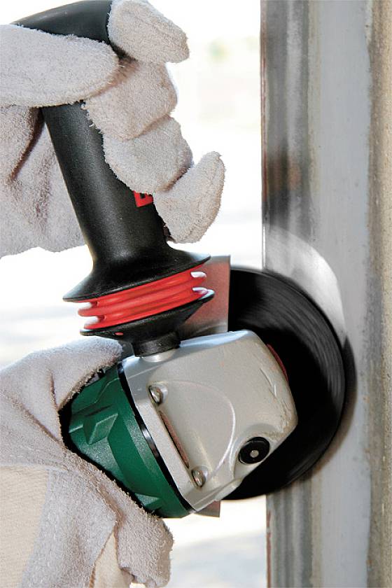 Gros plan sur une personne travaillant le bord d'une pièce métallique avec une meuleuse rotative. La personne porte des gants.