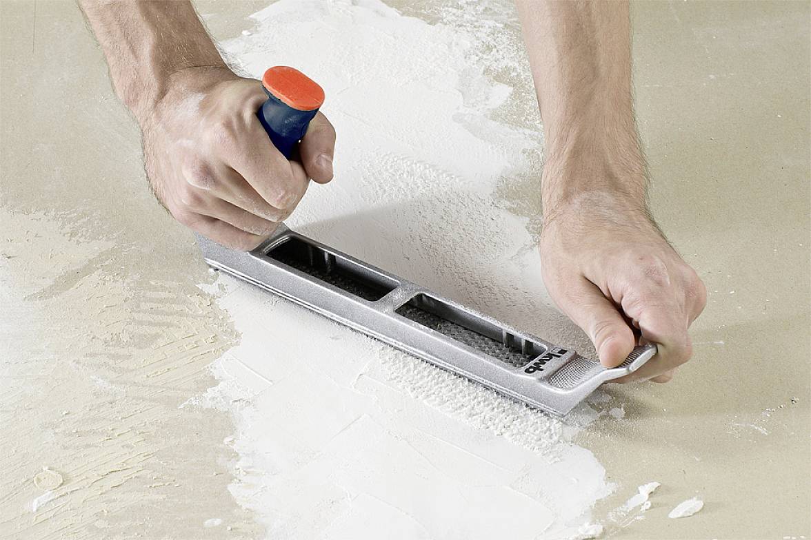 Des mains poncent une surface blanche à l'aide d'un outil de ponçage, éliminant le matériau excédentaire ou lissant la surface.