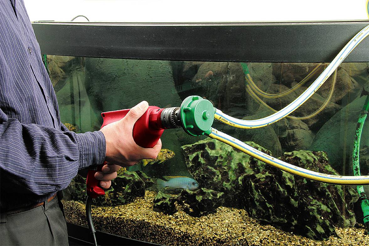 Une personne tient un outil rouge près d'un aquarium, avec des tuyaux plongeant dans l'eau. Un poisson nage en arrière-plan.