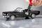 Une miniature d'une Ford Mustang Cabriolet noire est travaillée avec une perceuse rouge équipée d'un petit accessoire de meulage.
