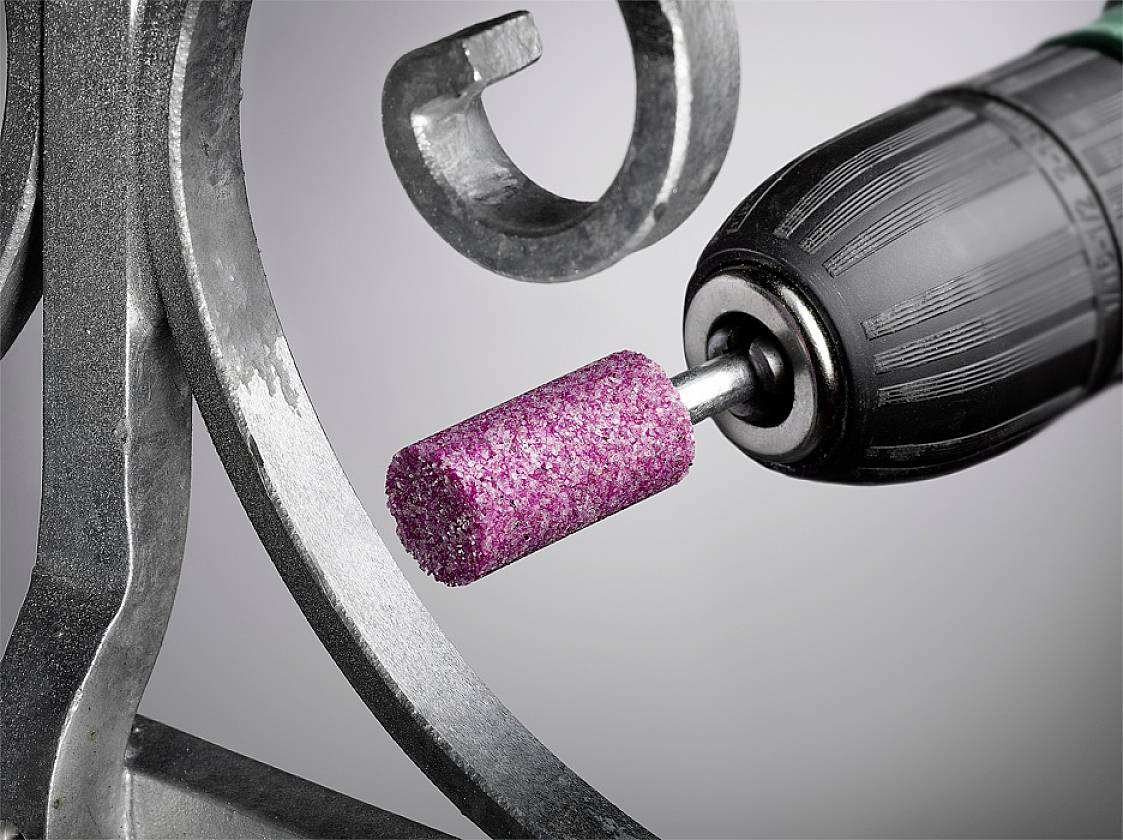 Une perceuse électrique avec une tête de meulage rose usiné une pièce métallique aux détails ondulés.