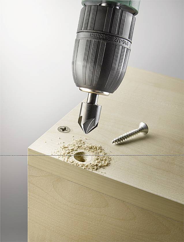 Une perceuse fore un large trou dans une planche de bois, à côté se trouve une tête de vis. Des copeaux de bois sont répartis autour du trou.