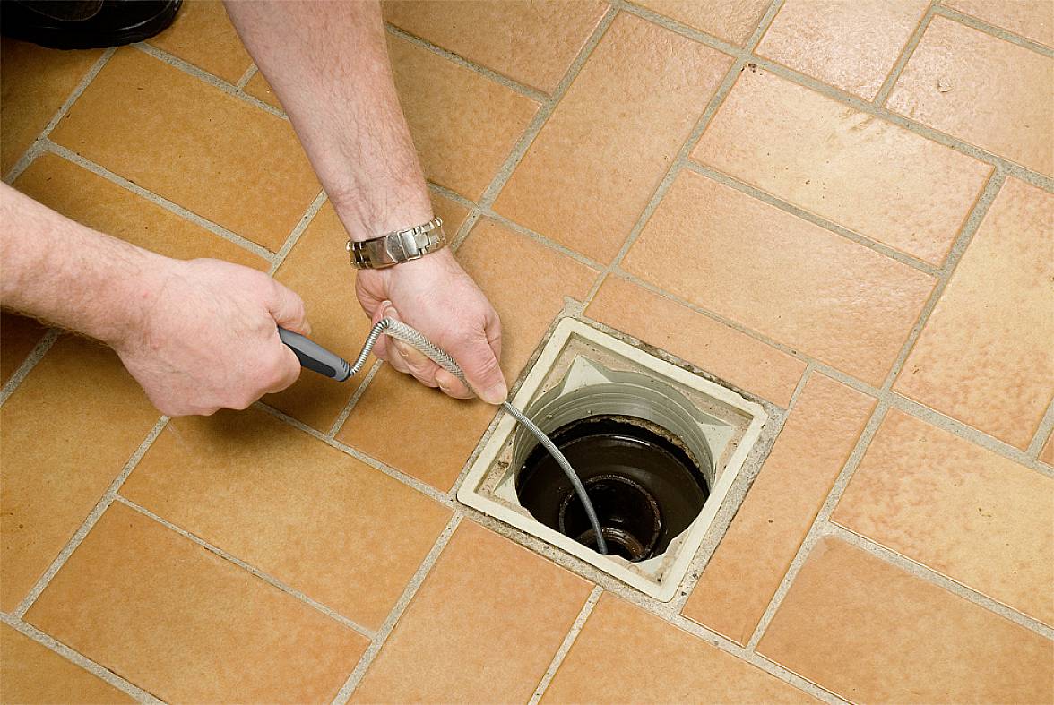 'Une personne nettoie un drain sur un sol carrelé à l'aide d'un outil de nettoyage flexible.'