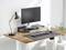 Bureau moderne avec surface en bois, écran, clavier, souris et lampe de bureau. Des plantes sont visibles en arrière-plan.