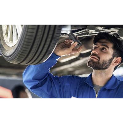 Un homme en tenue de travail bleue inspecte le soubassement d'une voiture à l'aide d'une lampe de poche dans un atelier.
