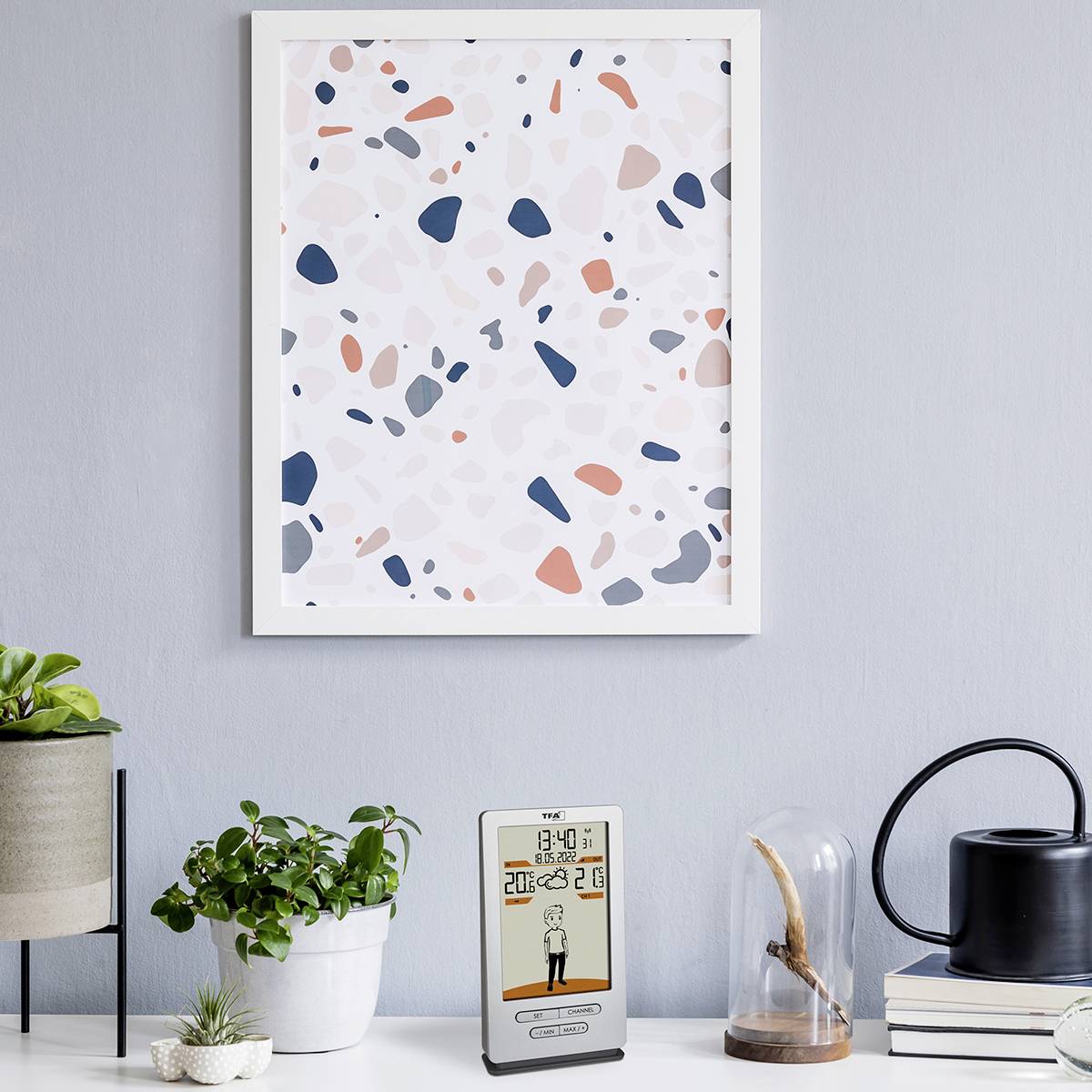 Un thermomètre numérique sur une table affiche '20,6°C'. À côté, des plantes vertes en pot et un tableau encadré sont accrochés au mur.