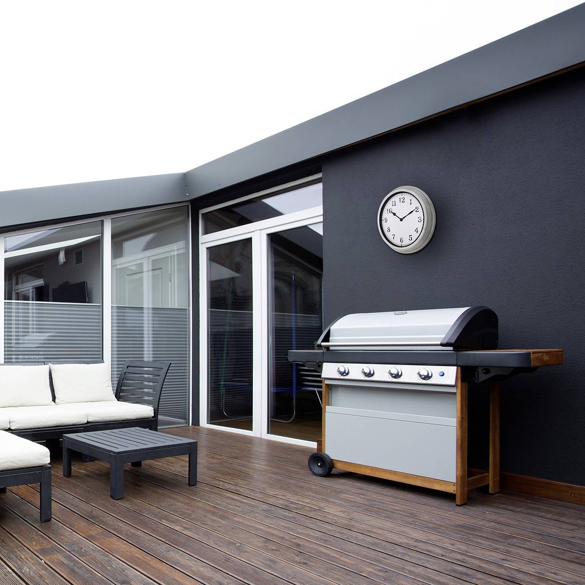 Terrasse avec un sol en bois, un canapé blanc et un barbecue moderne devant un mur de maison gris avec une horloge ronde.
