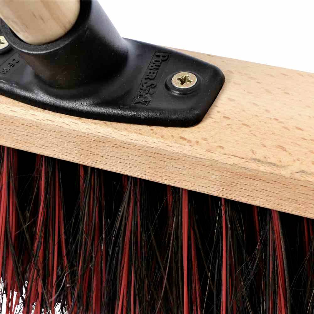 Un balai avec des poils rouges et noirs, fixé sur un manche en bois avec une attache métallique. Idéal pour balayer des saletés grossières.