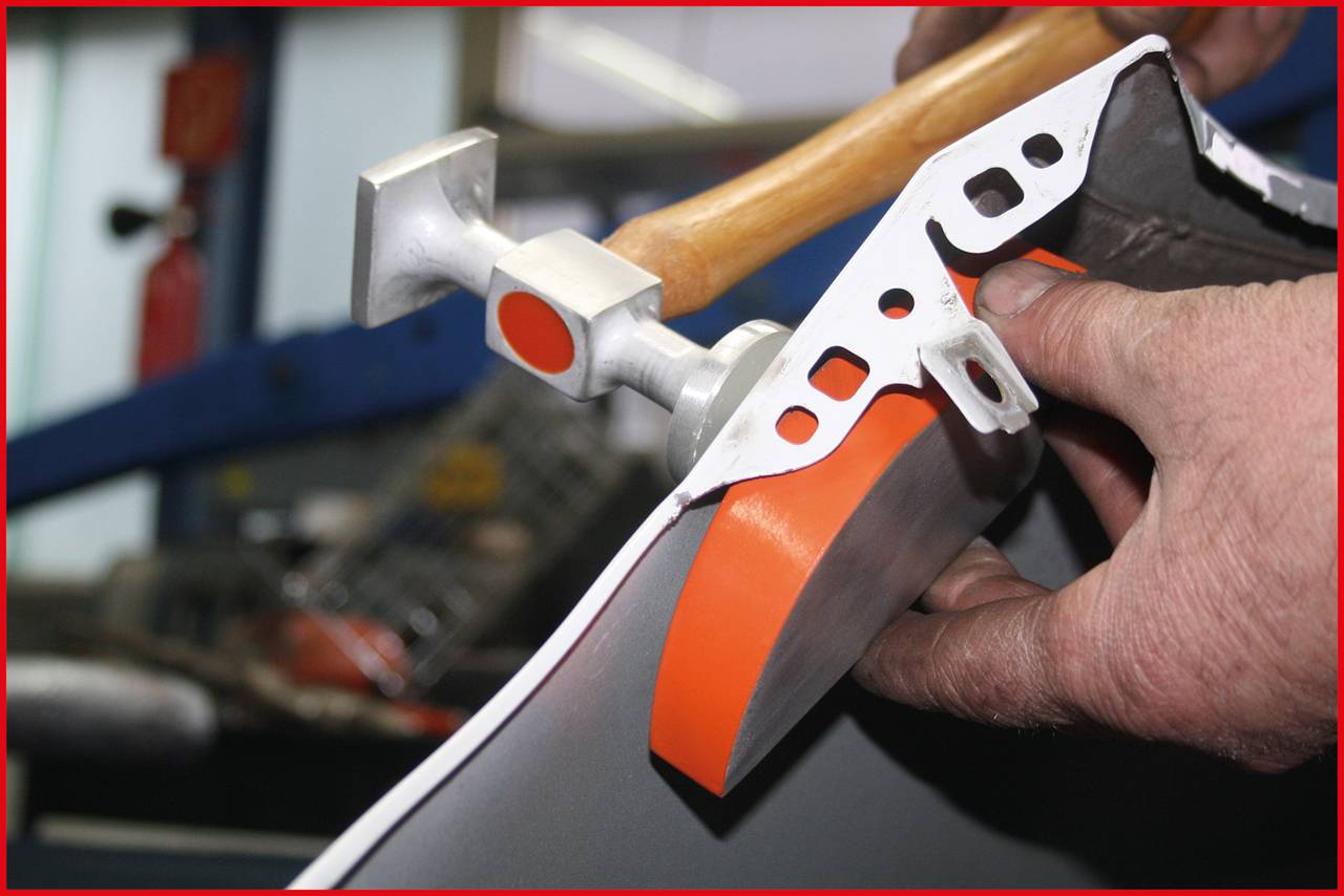 Une personne travaille un morceau de métal avec un marteau dans un atelier. L'accent est mis sur la partie d'usinage avec des détails orangés.