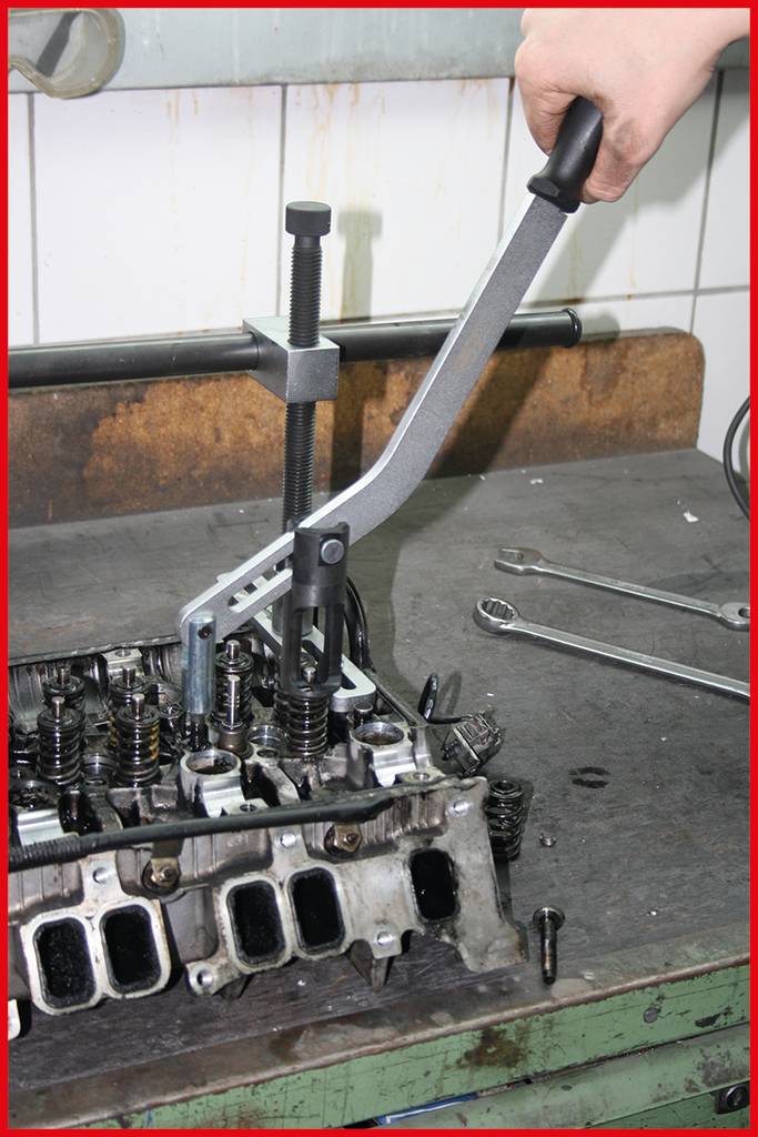 Une main utilise un outil pour tendre des ressorts de soupapes sur un bloc-moteur. Des clés sont posées sur l'établi en arrière-plan.
