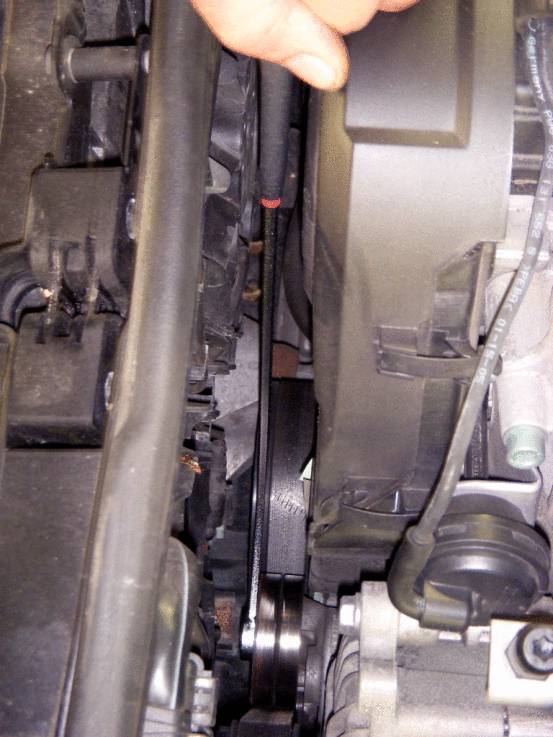La courroie trapézoïdale d'une voiture est visible, tandis qu'une main vient de dessus pour la vérifier. Des composants moteur sont disposés autour de la courroie.
