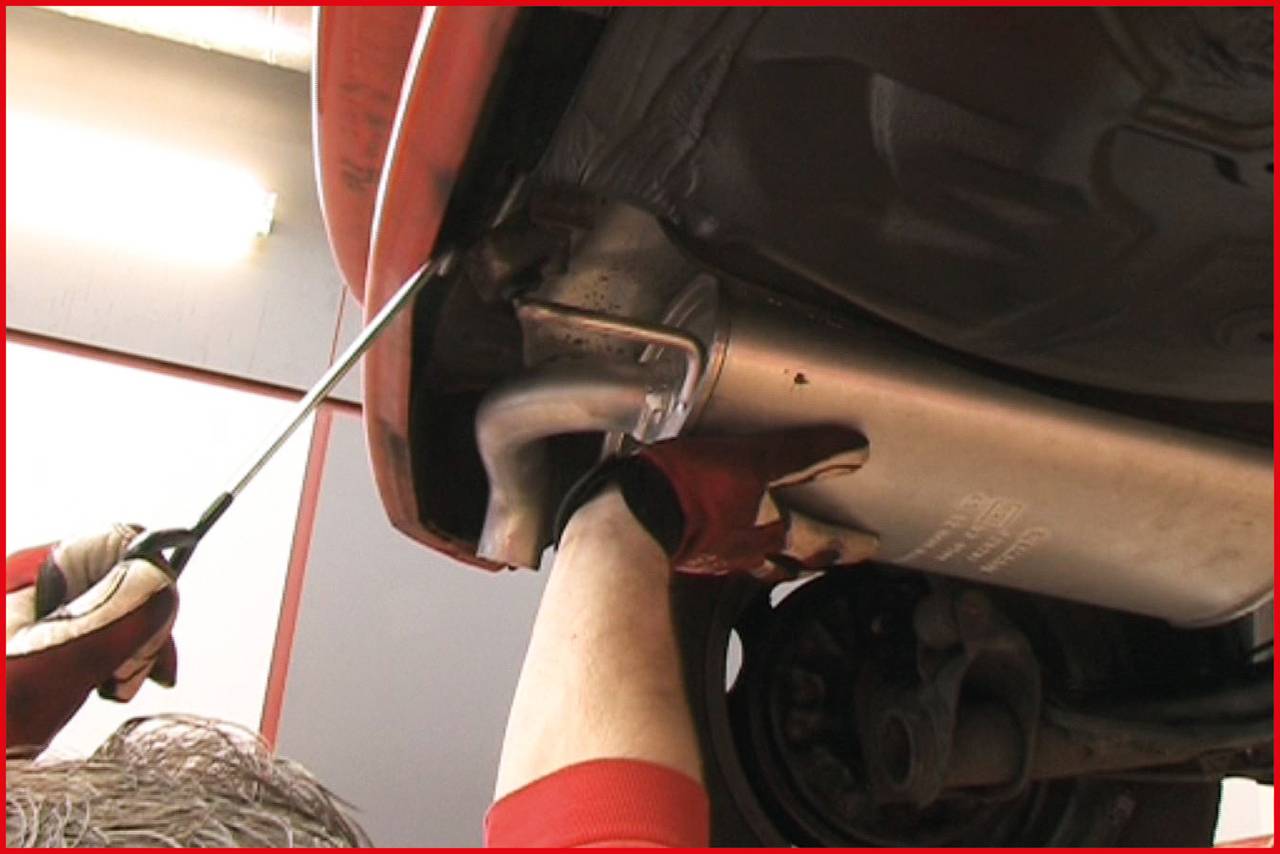 Une personne répare le pot d'échappement d'une voiture dans un atelier. Elle porte des gants et utilise un outil pour fixer le pot d'échappement.
