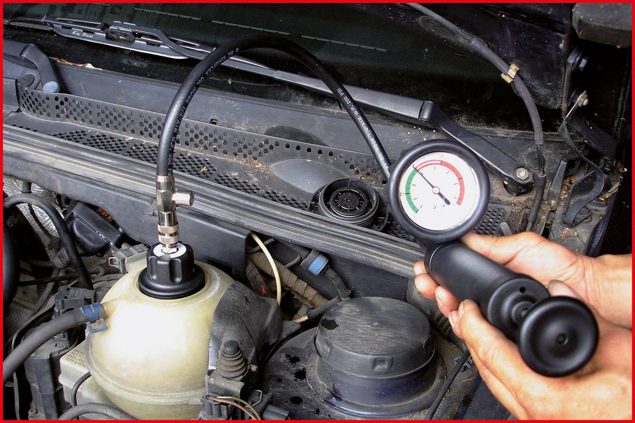 Appareil de mesure indiquant la pression dans le circuit de refroidissement d'une voiture. Des mains tiennent l'appareil, la pression se trouve dans la zone rouge. Compartiment moteur en arrière-plan.