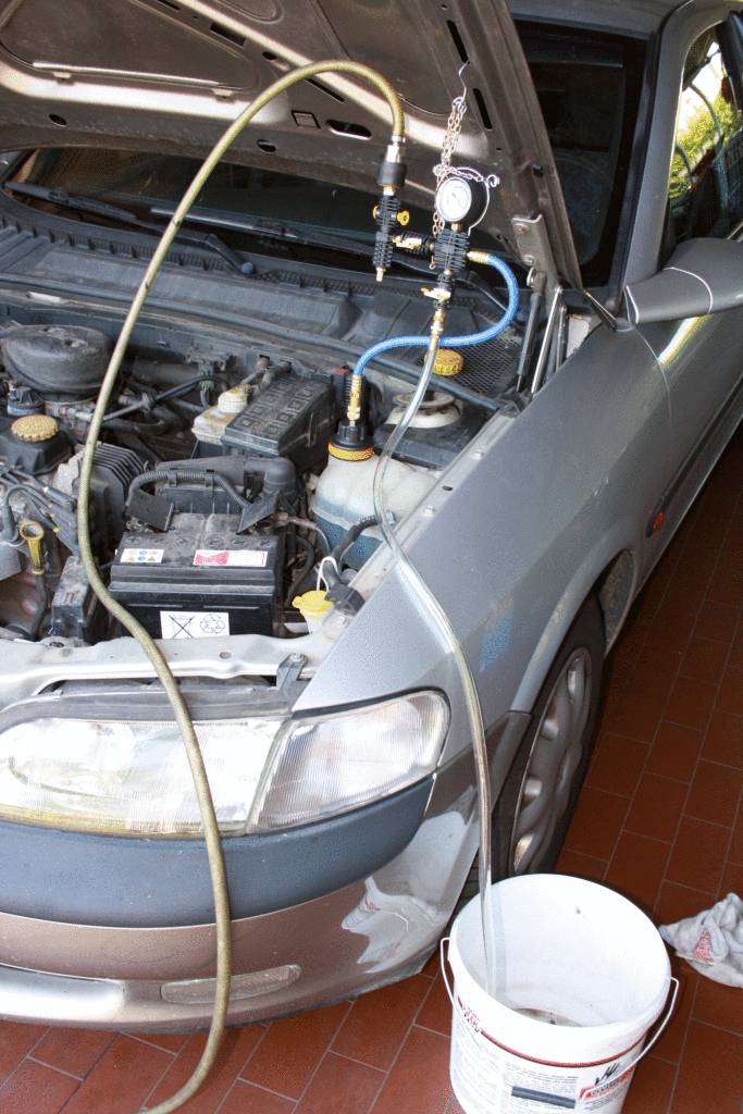 Une voiture avec le capot ouvert est en cours de ventilation. Des tuyaux et un manomètre sont branchés sur le moteur, un seau récupère le liquide.