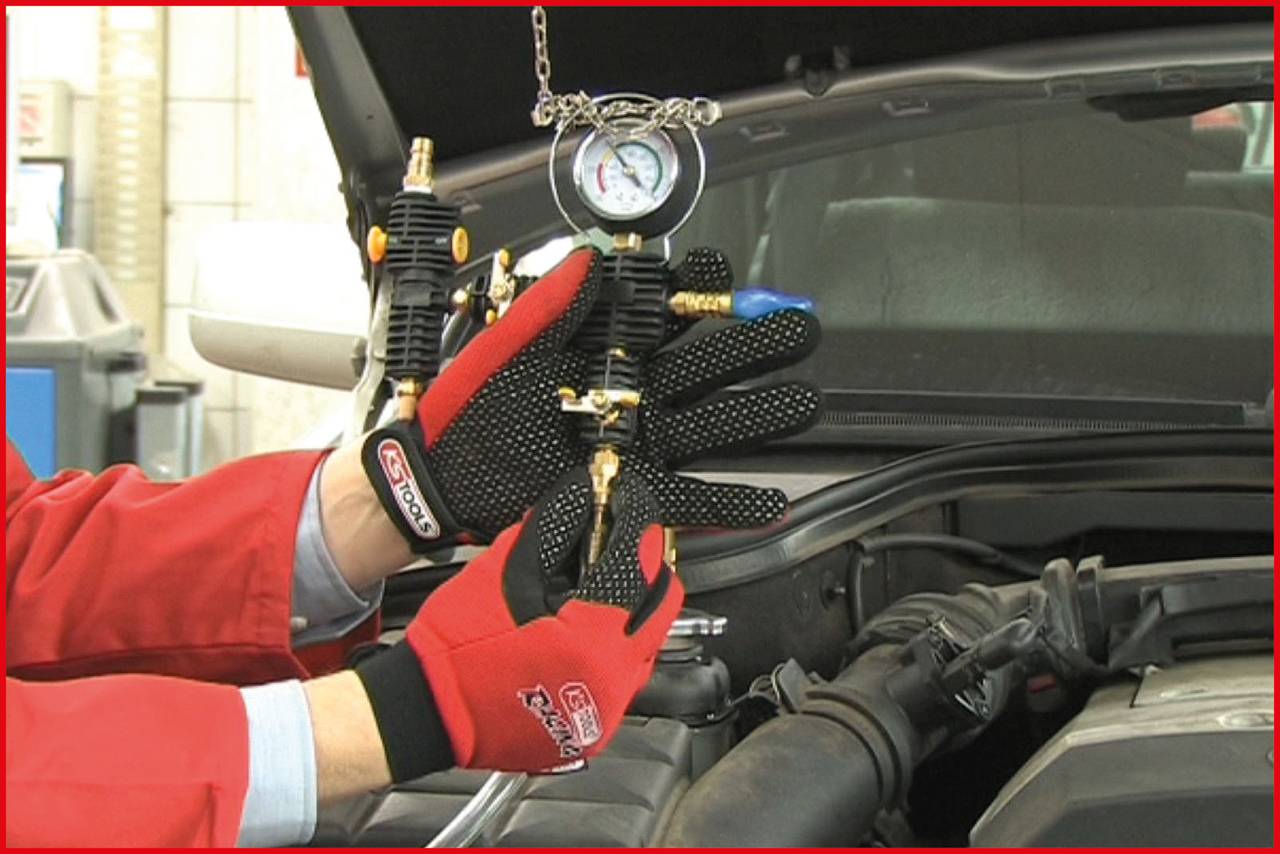 Une personne portant des gants rouges tient un manomètre près d'un compartiment moteur ouvert, probablement pour mesurer la pression.