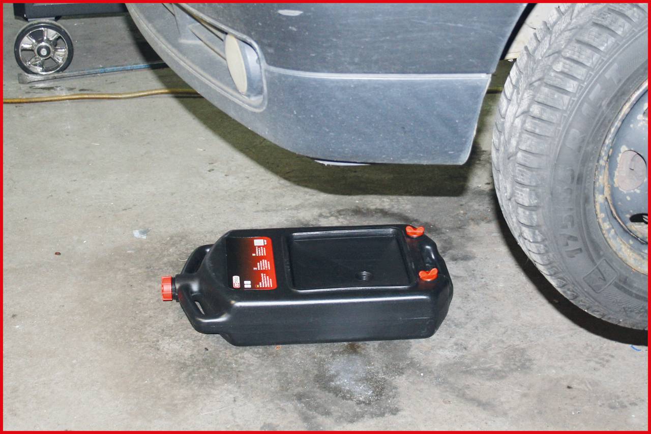 Un récipient noir de récupération d'huile avec un couvercle rouge est placé sous le compartiment moteur d'une voiture dans un garage. Un cric est visible à gauche.