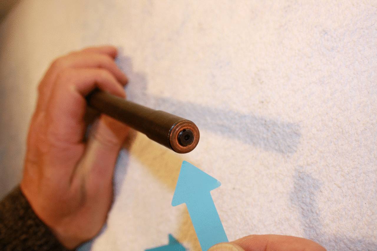 Une personne tient une tige brune contre un mur blanc, tandis qu'une autre main pointe une flèche bleue vers la zone circulaire de la tige.