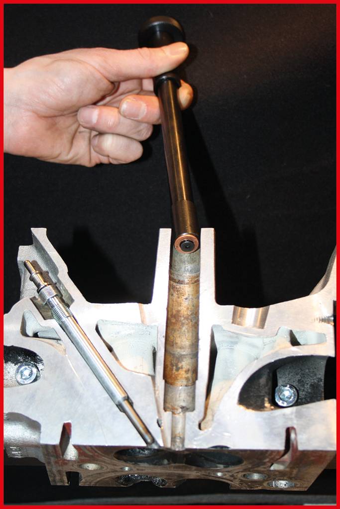 Une main tient une tige métallique au-dessus d'une pièce métallique usinée, qui pourrait être un composant de moteur.