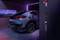 Une voiture électrique grise est en charge dans un garage futuriste. Des lumières néon illuminent la pièce et la station de recharge.