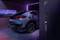 Une voiture électrique est garée dans un garage moderne et se charge sur une borne murale. Scène crépusculaire aux lumières violettes.