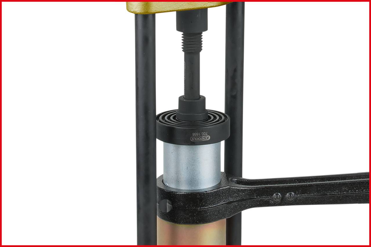 Une presse hydraulique comprime une pièce métallique cylindrique. Une tige métallique stabilise la pièce.