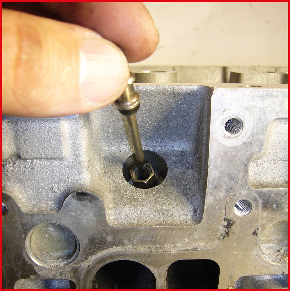Une main tient une petite soupape métallique qui est insérée dans une culasse de moteur. L'image montre un processus de réparation.