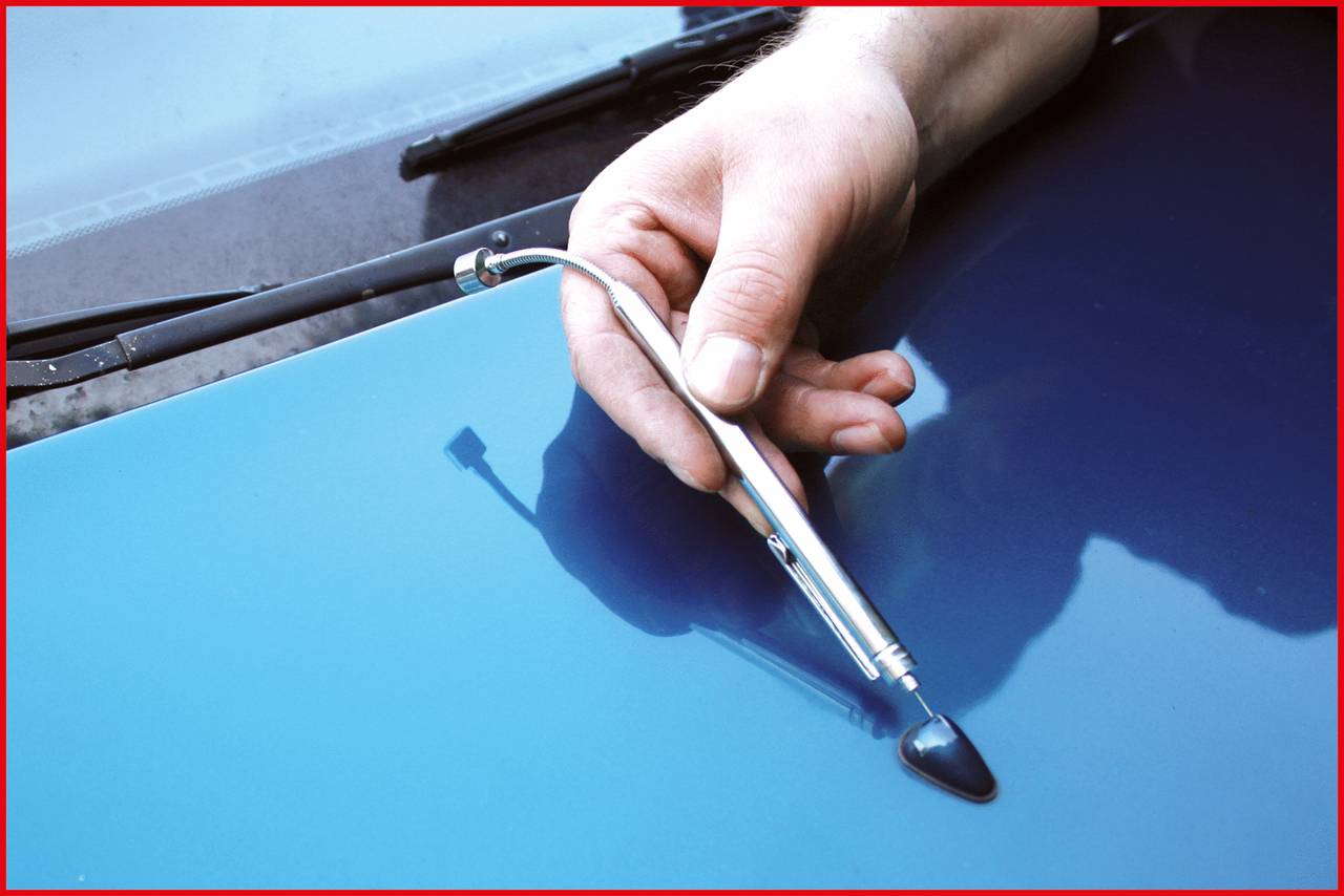 Une main tient un appareil de mesure d'épaisseur de peinture contre le capot bleu d'une voiture. L'appareil vérifie l'épaisseur de la couche de peinture.
