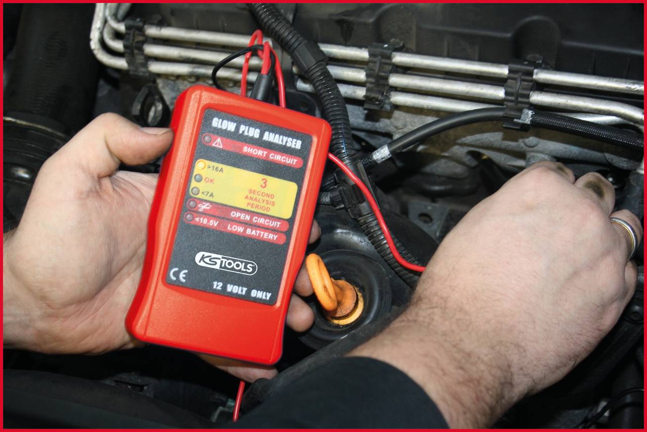 Une personne tient un dispositif de test de bougies de préchauffage au-dessus d'une pièce automobile. L'appareil affiche des valeurs de mesure. Des composants moteur sont visibles en arrière-plan.