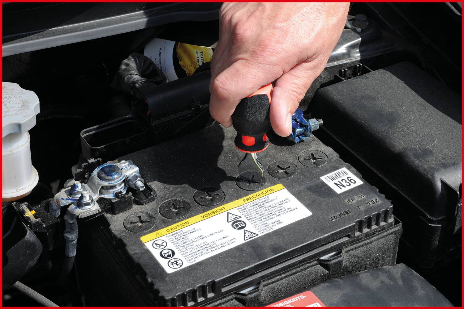Une main vérifie le niveau de liquide d'une batterie de voiture avec un tournevis sous le capot. Des avertissements sur la batterie sont visibles.
