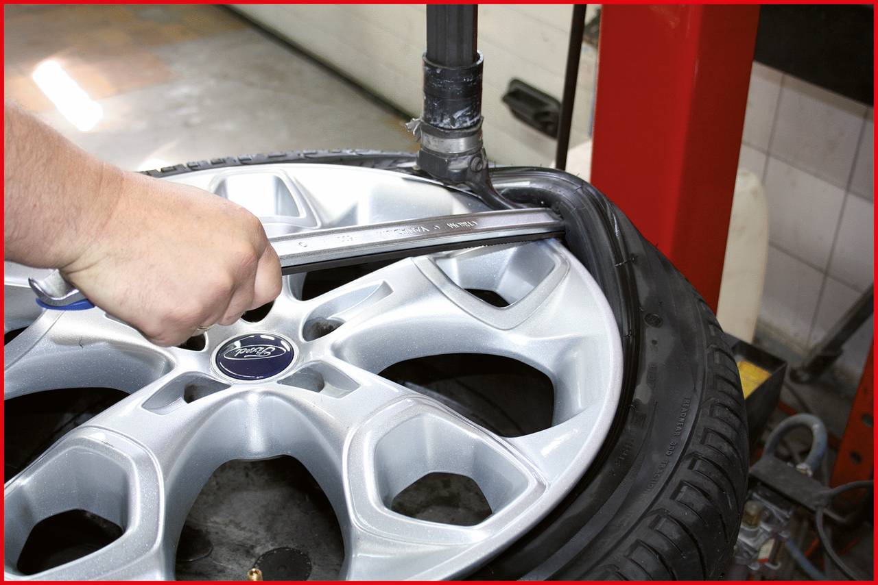 Une main utilise un outil pour monter un pneu sur une jante argentée portant le logo Ford. Scène dans un atelier.
