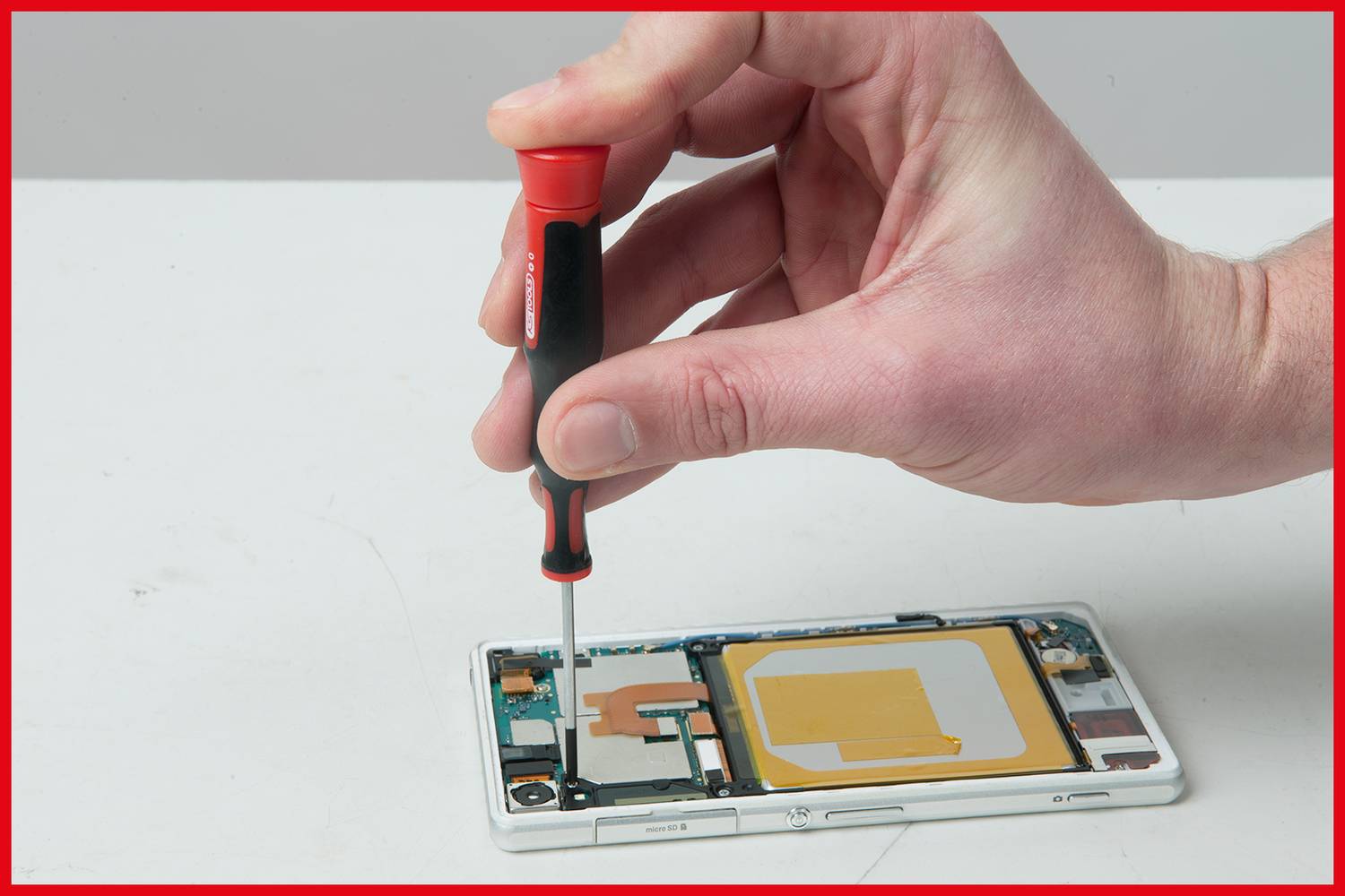 Une main tient un tournevis et ouvre le dos d'un smartphone. Des composants internes sont visibles, notamment la batterie et la carte mère.