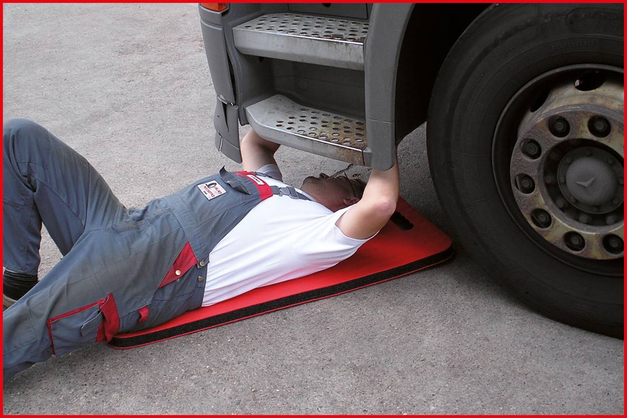 Une personne en tenue de travail est allongée sous un camion, apparemment en train d'effectuer des réparations, sur un tapis rembourré.