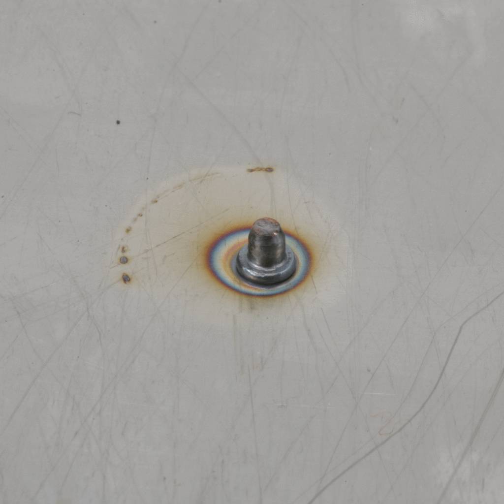 Un boulon est soudé à une surface métallique, entouré d'une zone légèrement brûlée suite au travail de soudure.