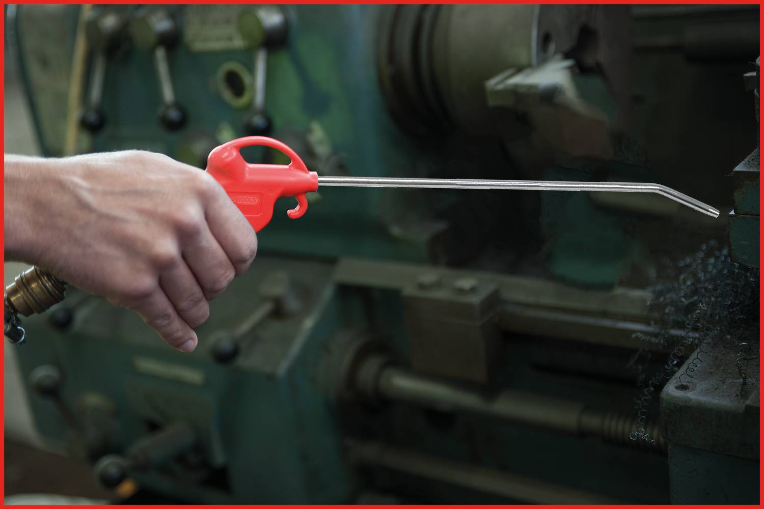 Une personne tient une bombe à air comprimé avec une poignée rouge pour enlever des copeaux métalliques d'une machine. La machine est partiellement visible.