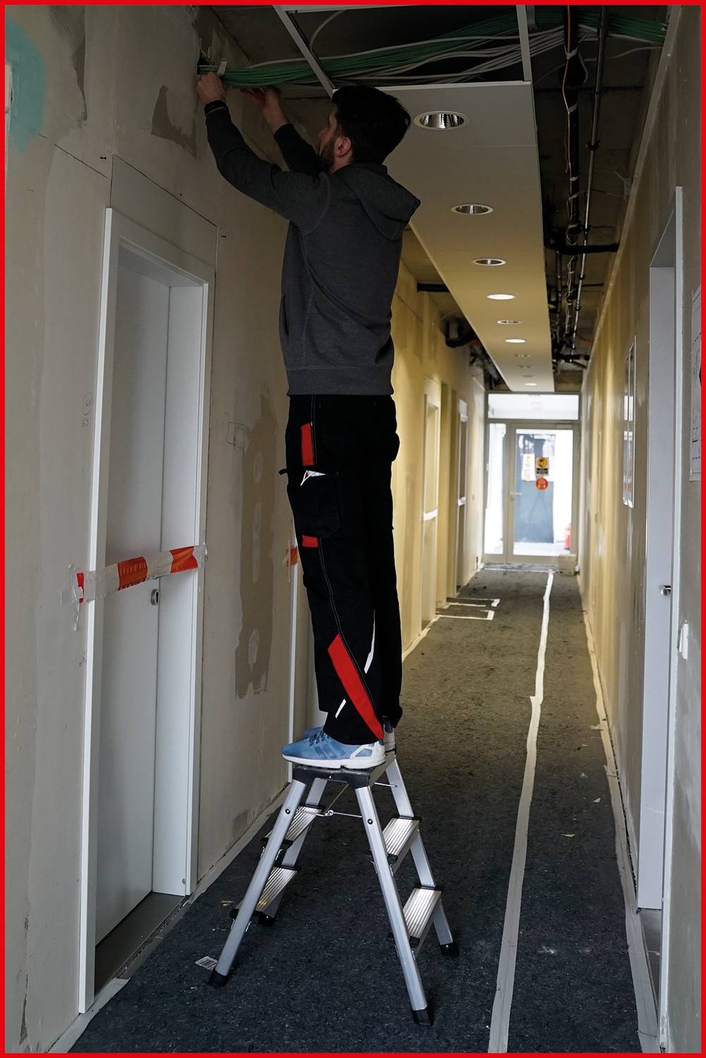 Un homme est debout sur une échelle et travaille au plafond dans un long couloir. Certaines parties du mur sont enduites de manière incomplète.