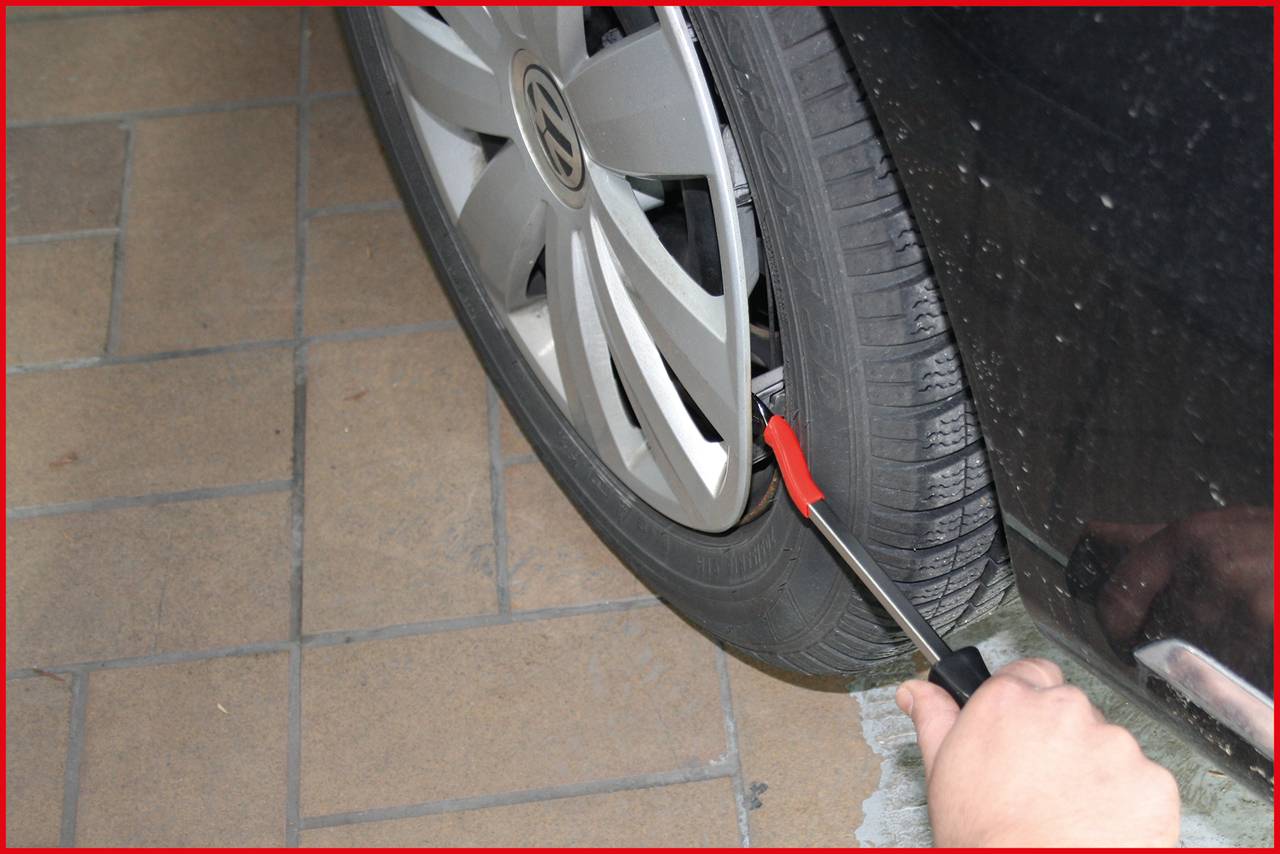 Une main tient un outil qui est inséré dans la valve d'un pneu de voiture pour vérifier la pression. La voiture est garée sur un sol pavé.