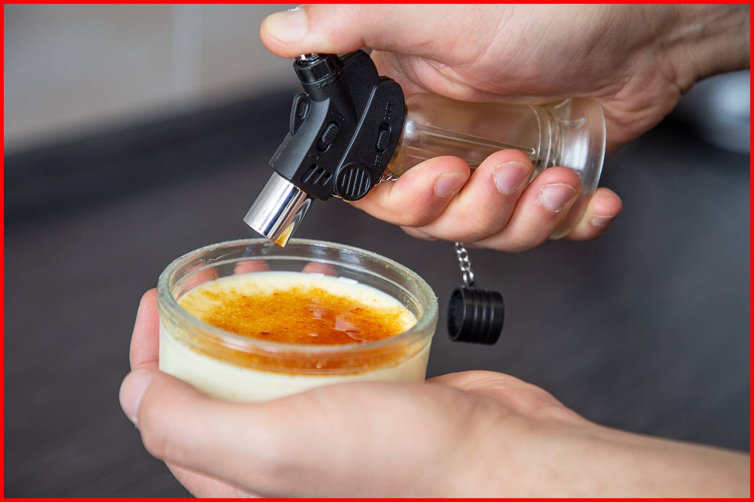 Une main tient un petit chalumeau de pâtisserie au-dessus d'une crème brûlée pour caraméliser la surface de sucre.