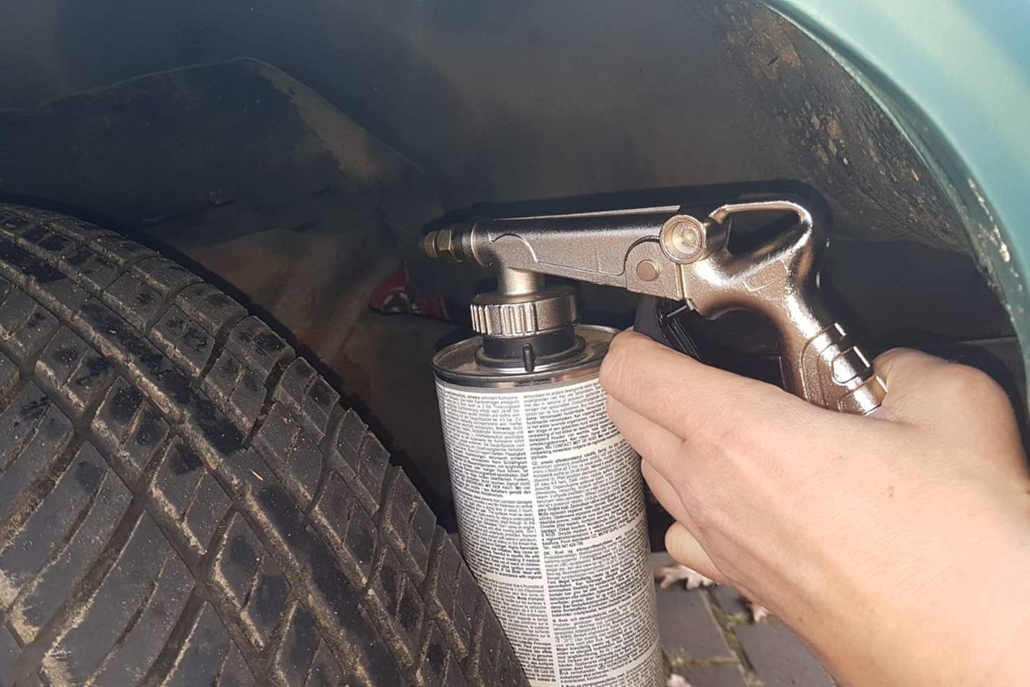 Gros plan sur une main appliquant un aérosol sur la suspension de roue d'une voiture. La voiture est de couleur turquoise, et le pneu est visible.