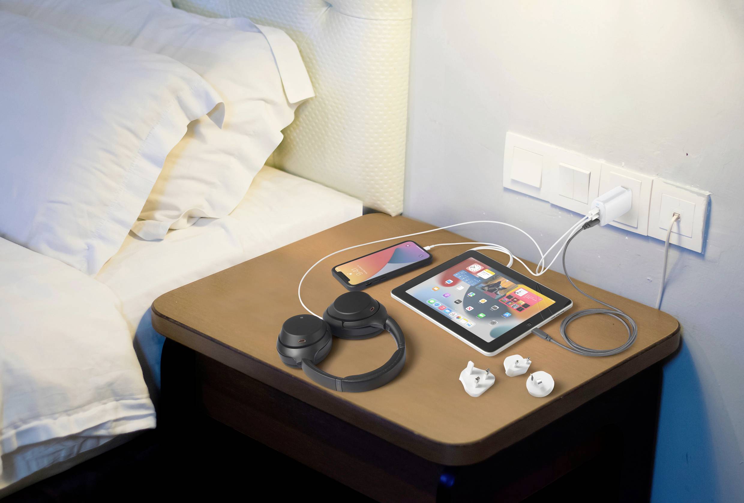 Table de nuit avec des écouteurs, une tablette, un smartphone et un chargeur. La tablette affiche des applications. Deux prises électriques sont fixées au mur.