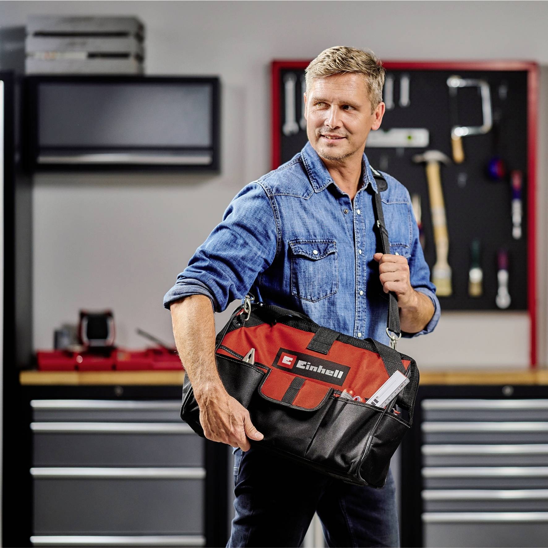 Un homme en tenue de travail tient un sac à outils devant un mur d'atelier garni de divers outils et tiroirs.