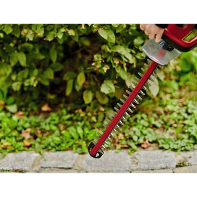 Une personne tient un taille-haie rouge devant un buisson vert. Le taille-haie est prêt à l'emploi et sera utilisé pour couper des branches.